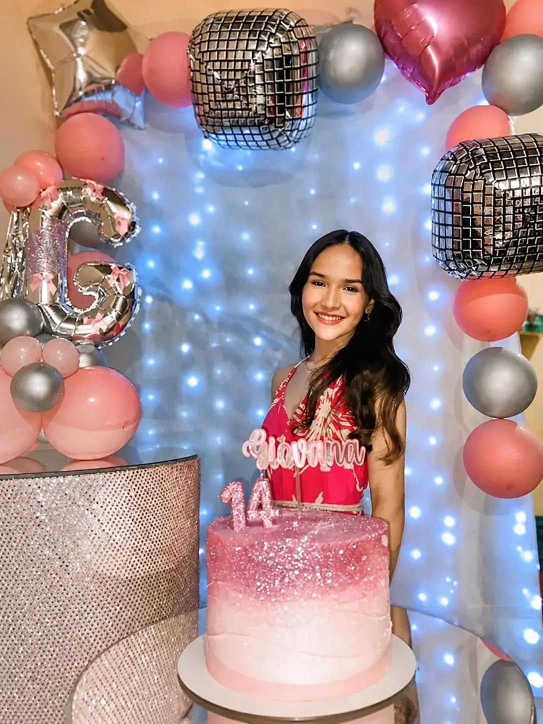 Jovem sorrindo ao lado de um bolo rosa e de balões, na comemoração de 14 anos