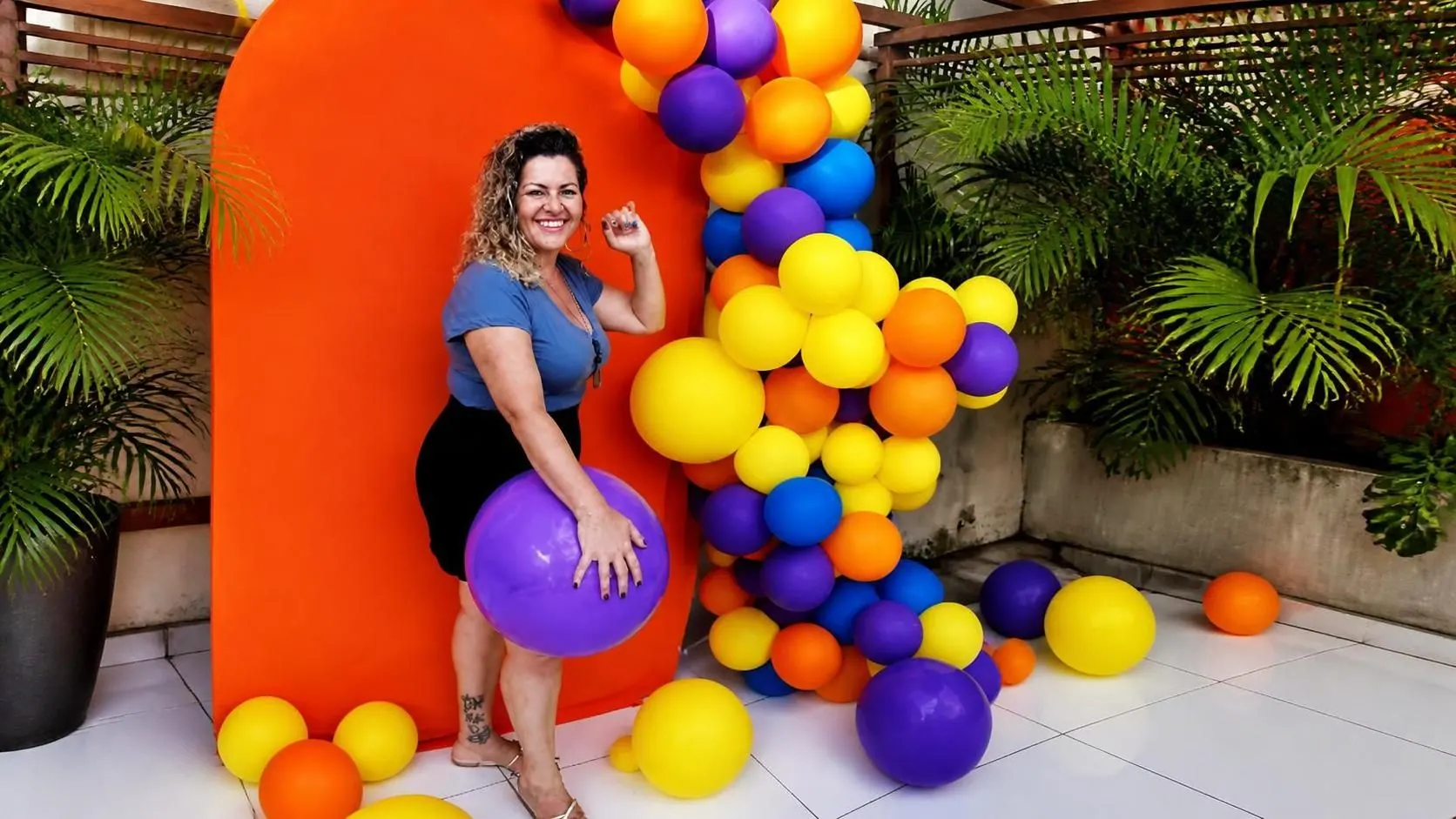 Mulher sorrindo ao lado de um arco de balões laranja, roxos, azuis e amarelos